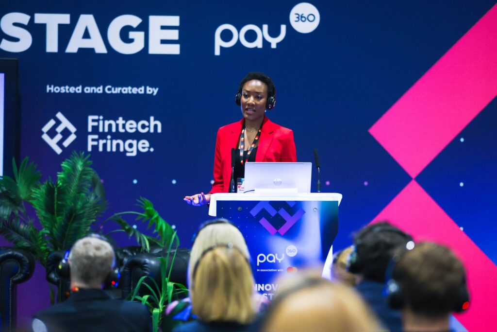 Nkechi, in a bright red jacket, presents from a podium. Pay360 event branding is prominently in the background.