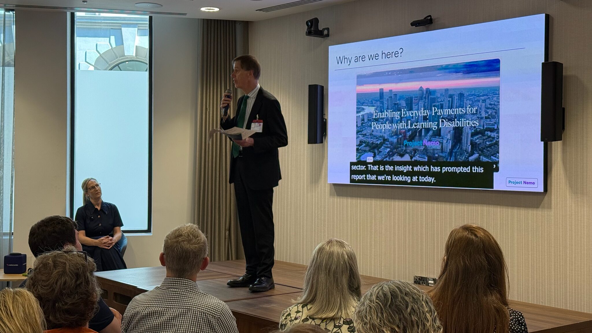 A middle-aged man in a suit speaks to an audience in an event space. Behind him, a screen reads "enabling everyday payments for people with learning disabilities"
